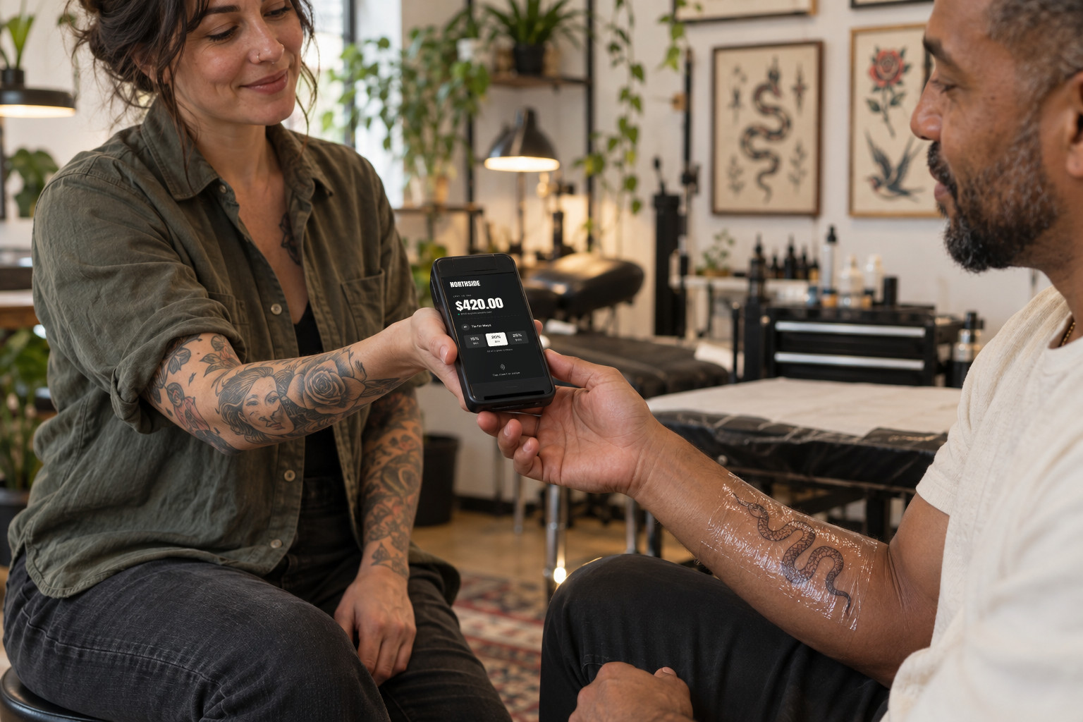 An artist hands a card terminal showing the Studio's own tip screen to a client after a session
